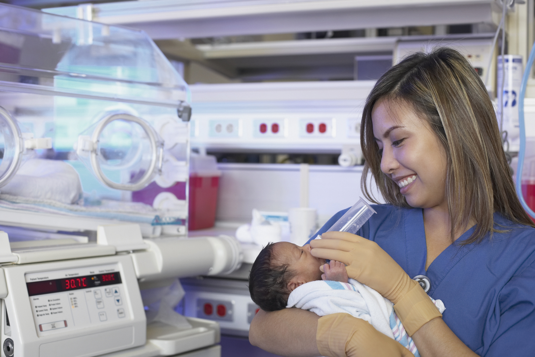 Female nurse feeding newborn baby next to incubator